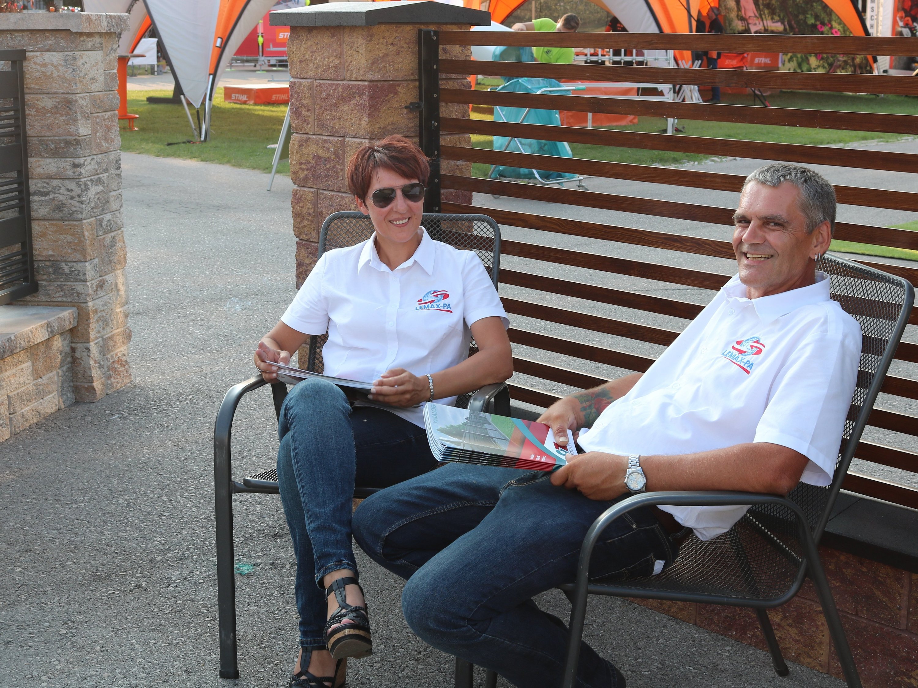 Two Lemax PA employees sit relaxed on garden chairs, smiling and holding brochures - personal advice with heart.